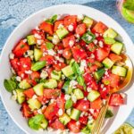 overhead flat lay of watermelon feta salad in a large white serving bowl with fresh mint, crumbled feta, sliced cucumber, and a honey-lime drizzle on a bright summer table