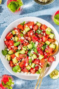 overhead flat lay of watermelon feta salad in a large white serving bowl with fresh mint, crumbled feta, sliced cucumber, and a honey-lime drizzle on a bright summer table