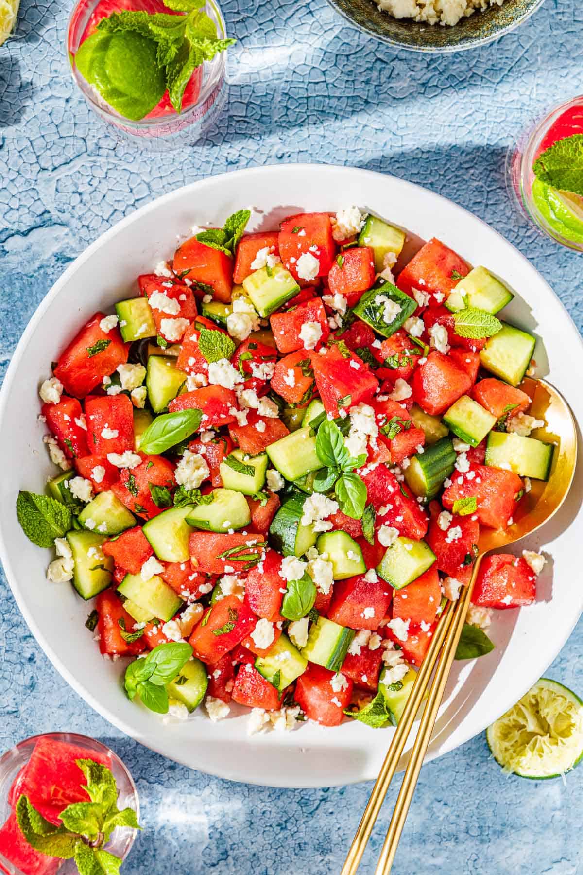 overhead flat lay of watermelon feta salad in a large white serving bowl with fresh mint, crumbled feta, sliced cucumber, and a honey-lime drizzle on a bright summer table