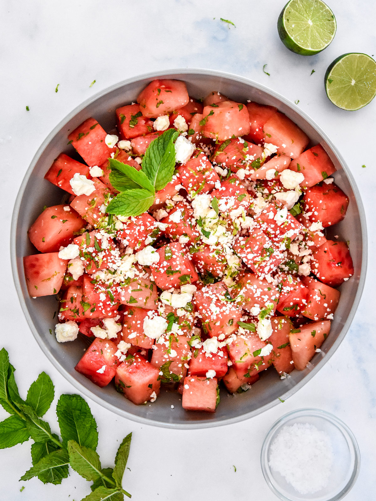 flat lay ingredients shot showing cubed watermelon, block of feta, fresh mint leaves, cucumber slices, lime, honey, and olive oil arranged on a marble surface