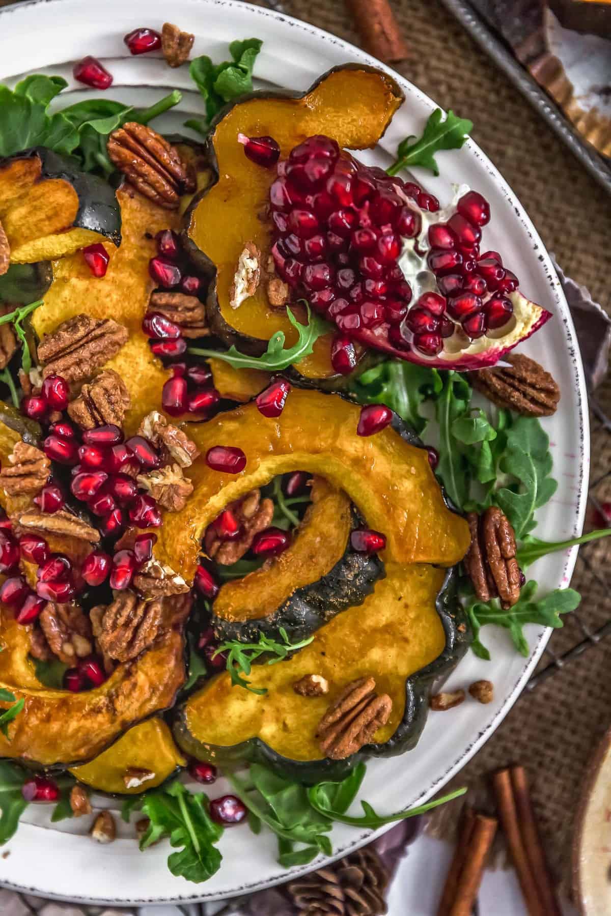 Acorn squash wedges being tossed in a maple-cinnamon glaze in a large mixing bowl, with cinnamon sticks, maple syrup bottle, and olive oil on the counter