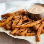 Close-up of a single crispy butternut squash fry being dipped into a creamy Greek-yogurt chipotle sauce in a small white bowl, with lime wedges and fresh herbs in the background