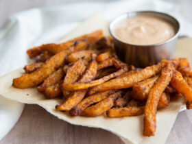 Air Fryer Butternut Squash Fries (Crispy, Healthy & Ready in 25 Minutes) 3 Close-up of a single crispy butternut squash fry being dipped into a creamy Greek-yogurt chipotle sauce in a small white bowl, with lime wedges and fresh herbs in the background