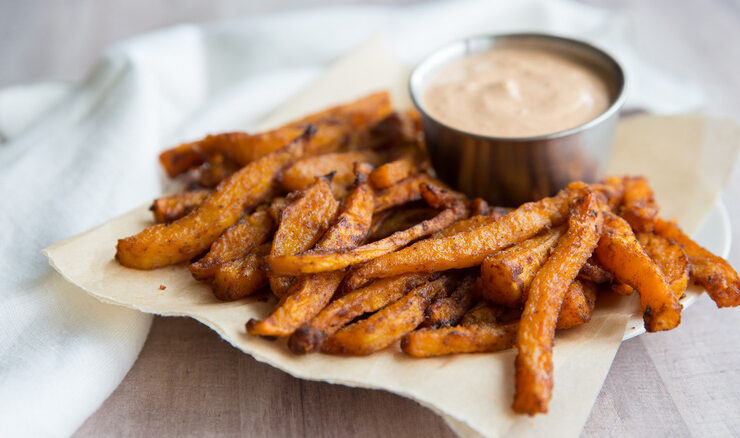 Close-up of a single crispy butternut squash fry being dipped into a creamy Greek-yogurt chipotle sauce in a small white bowl, with lime wedges and fresh herbs in the background