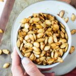 Bowl of golden crispy air fryer pumpkin seeds seasoned with sea salt and cinnamon, photographed overhead on a wooden surface with a small carved pumpkin in the background