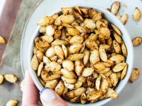 Bowl of golden crispy air fryer pumpkin seeds seasoned with sea salt and cinnamon, photographed overhead on a wooden surface with a small carved pumpkin in the background