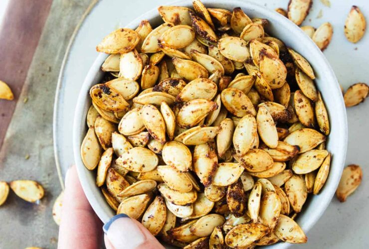 Bowl of golden crispy air fryer pumpkin seeds seasoned with sea salt and cinnamon, photographed overhead on a wooden surface with a small carved pumpkin in the background