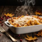 Baked ziti in a white ceramic casserole dish, bubbling with golden-brown mozzarella cheese on top, fresh basil garnish, rustic wooden table setting