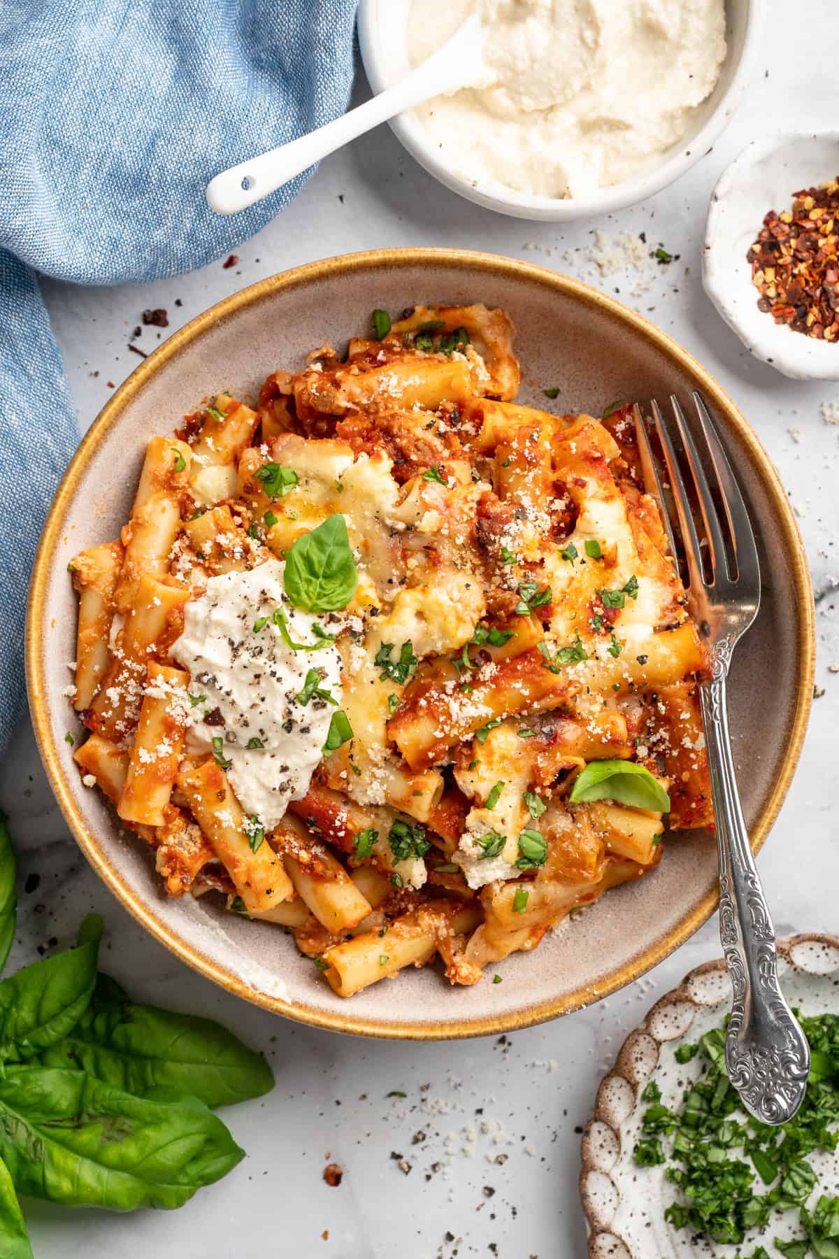 Overhead flat-lay of baked ziti ingredients — ziti pasta, ricotta in a bowl, shredded mozzarella, ground beef, jar of marinara sauce, fresh herbs on a light marble surface