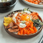 A beautifully plated Bibimbap Korean rice bowl with colorful sautéed vegetables, a sunny-side-up egg, and a drizzle of gochujang sauce, served in a stone bowl