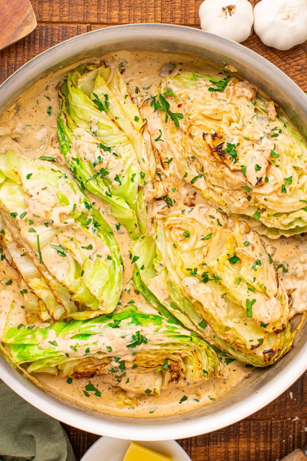 Overhead flat-lay of halved green cabbage, a block of Parmigiano-Reggiano, a small bowl of heavy cream, whole garlic cloves, a stick of butter, and a bottle of avocado oil arranged on a white marble surface