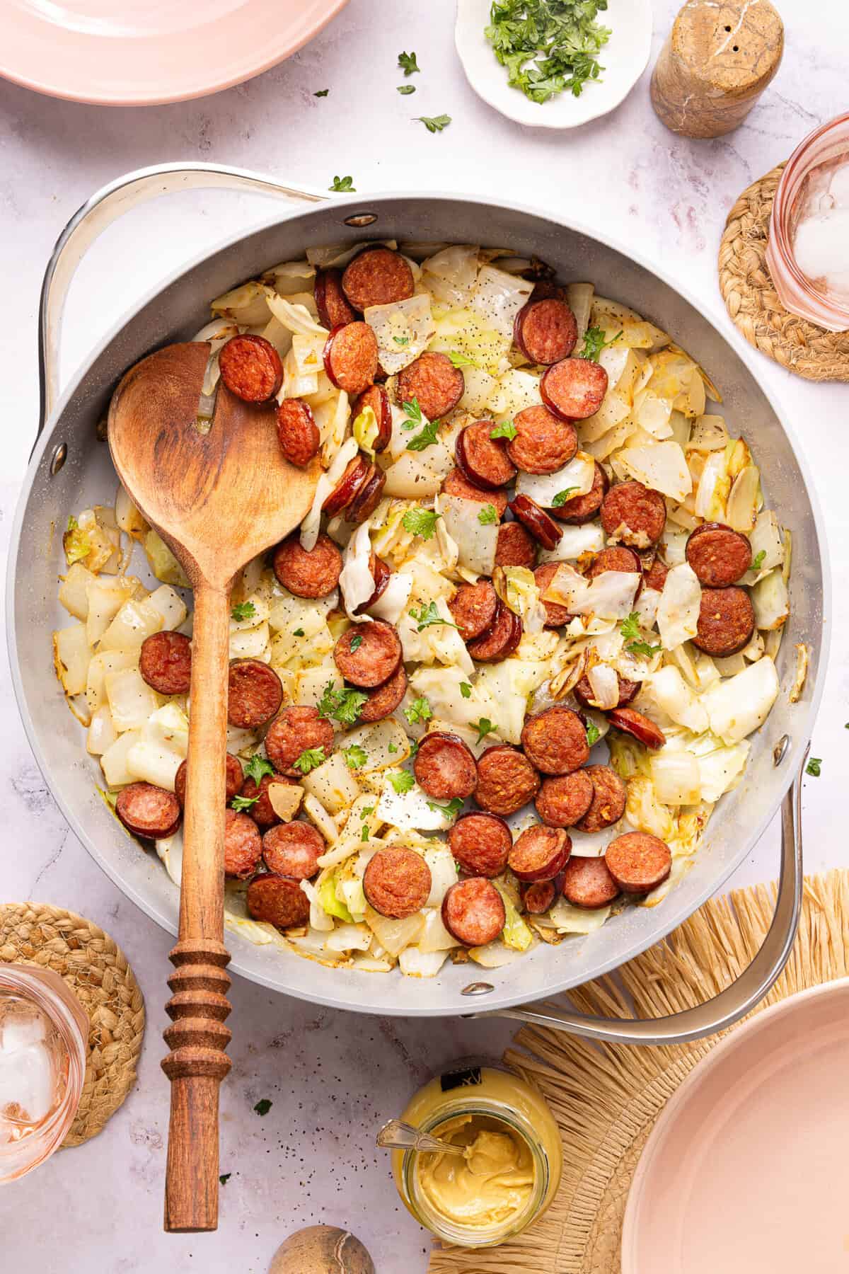 Overhead flat-lay of cabbage and sausage skillet ingredients — sliced kielbasa, shredded cabbage, halved onion, garlic cloves, paprika, and apple cider vinegar on a wooden board