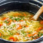 A steaming bowl of from-scratch crockpot chicken noodle soup with golden broth, tender shredded chicken, egg noodles, and fresh parsley, photographed on a rustic wooden table