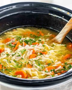 A steaming bowl of from-scratch crockpot chicken noodle soup with golden broth, tender shredded chicken, egg noodles, and fresh parsley, photographed on a rustic wooden table