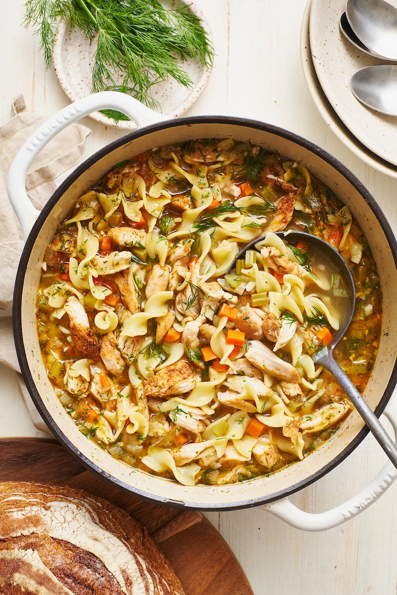 Overhead flat-lay of crockpot chicken noodle soup ingredients — bone-in chicken pieces, chopped carrots, celery, onion, garlic, fresh herbs, and dry egg noodles arranged on a white marble surface
