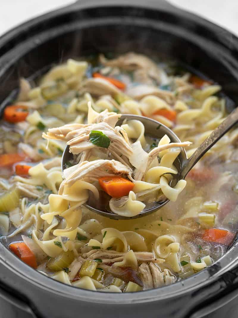 Close-up overhead shot of a ladle lifting a generous scoop of crockpot chicken noodle soup from the slow cooker, showing shredded chicken, egg noodles, and vegetable-studded golden broth