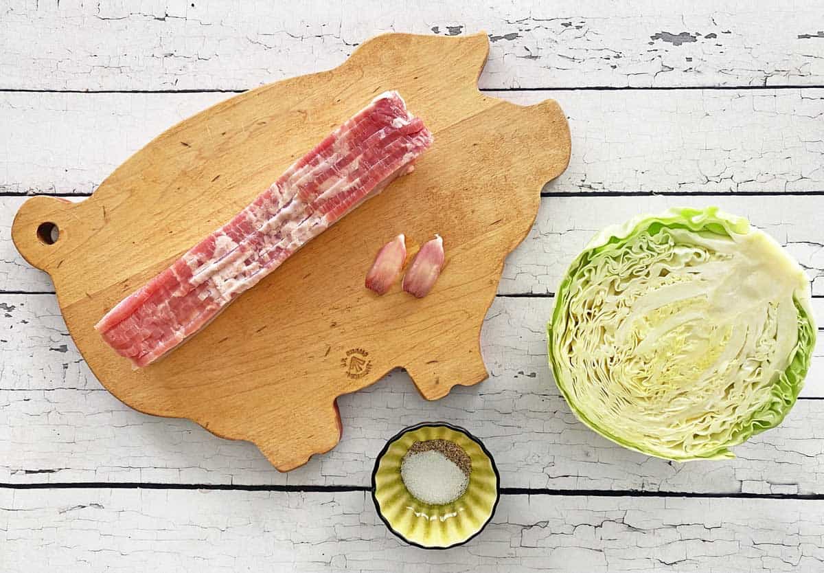 Overhead flat lay of raw cabbage wedges, strips of thick-cut bacon, yellow onion, garlic cloves, apple cider vinegar bottle, and black pepper on a wooden cutting board