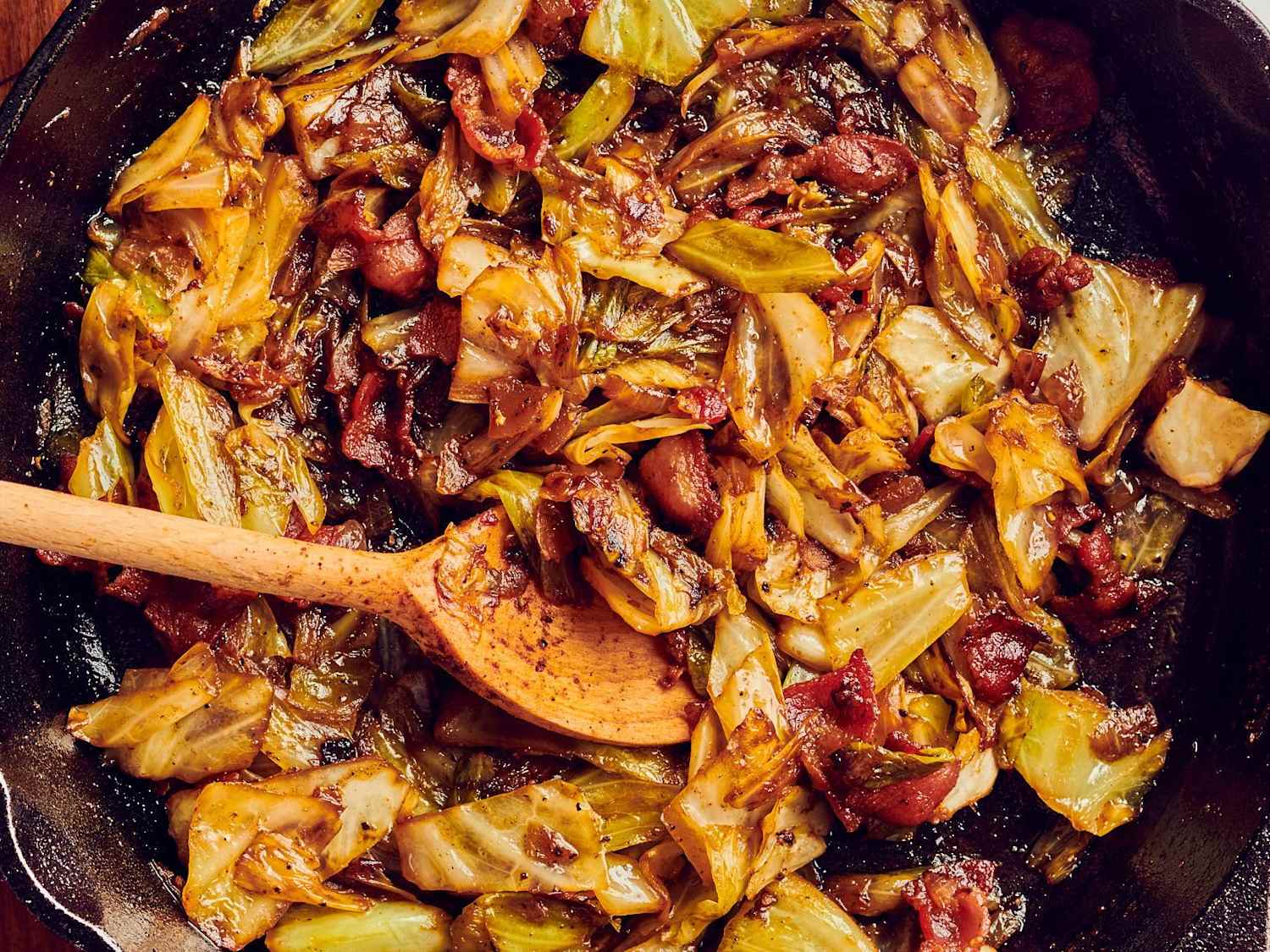 Close-up of golden fried cabbage with crispy bacon pieces in a cast iron skillet, showing caramelized edges and steam rising, photographed from slightly above