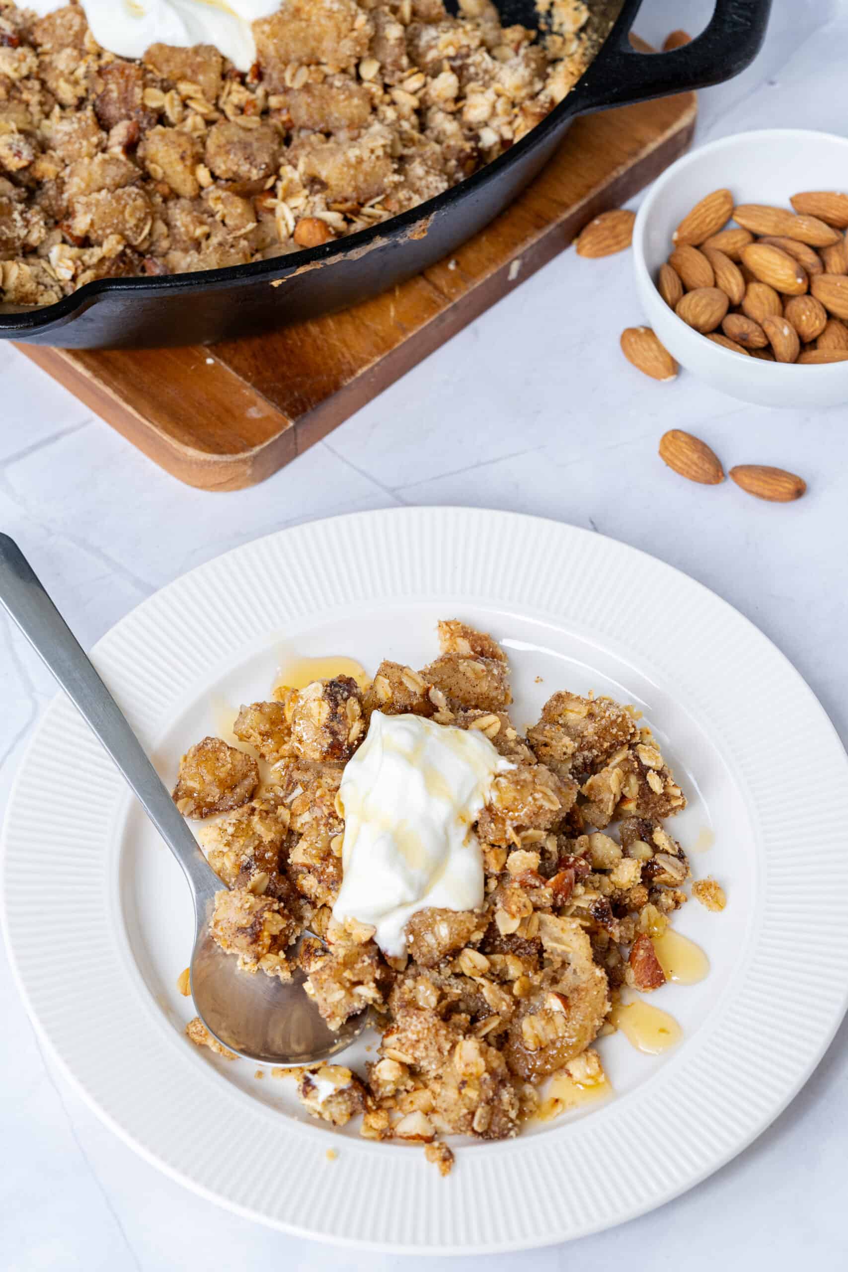 Greek yogurt apple crisp in a white ceramic baking dish, golden oat-almond streusel on top with a generous swirl of thick Greek yogurt and a sprinkle of cinnamon, shot overhead on a warm wooden surface with a linen napkin and whole apples nearby