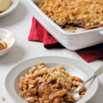 Flat-lay of all key ingredients for greek yogurt apple crisp — sliced apples, rolled oats, sliced almonds, butter cubes, cinnamon sticks, maple syrup, and a bowl of thick Greek yogurt on a marble surface
