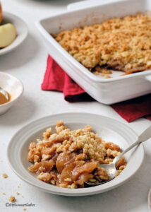 Flat-lay of all key ingredients for greek yogurt apple crisp — sliced apples, rolled oats, sliced almonds, butter cubes, cinnamon sticks, maple syrup, and a bowl of thick Greek yogurt on a marble surface