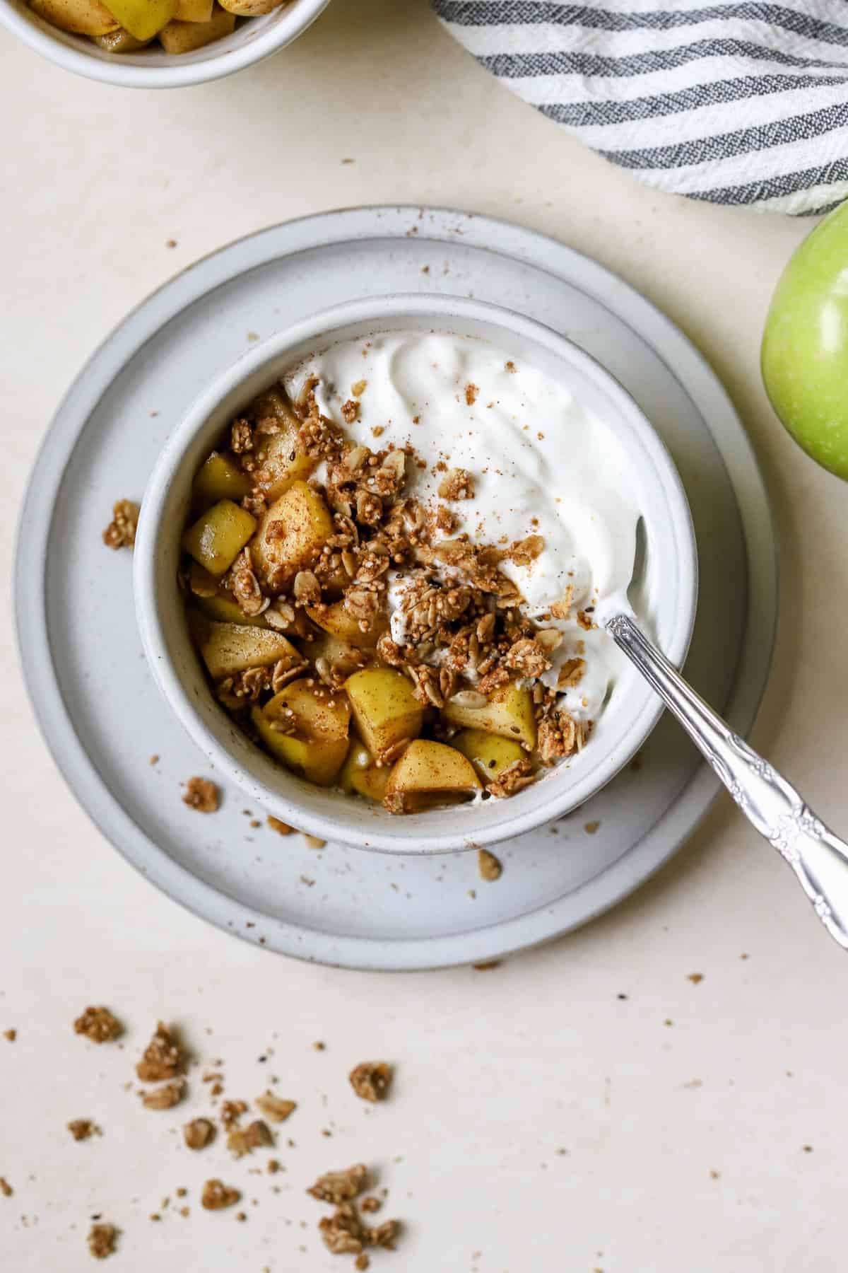 Close-up of a single serving of greek yogurt apple crisp in a white bowl, showing layers of caramel-soft apple slices beneath a craggy golden oat-and-almond streusel, topped with a generous cold swirl of Greek yogurt and a dusting of cinnamon, with a spoon resting alongside