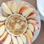 Greek yogurt caramel apple dip in a white bowl with a caramel swirl on top, surrounded by crisp apple slices on a wooden board