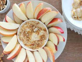 Greek yogurt caramel apple dip in a white bowl with a caramel swirl on top, surrounded by crisp apple slices on a wooden board