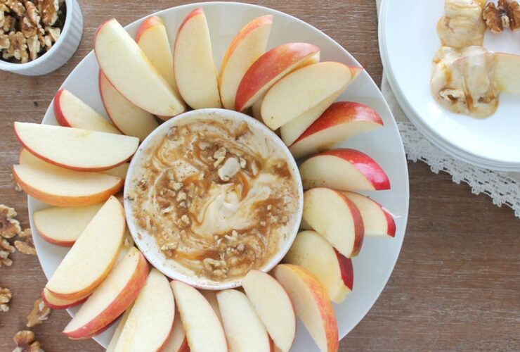 Greek yogurt caramel apple dip in a white bowl with a caramel swirl on top, surrounded by crisp apple slices on a wooden board