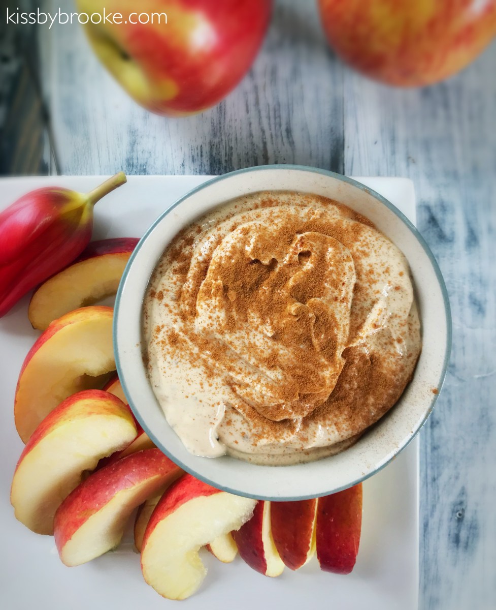 Overhead flat-lay of Greek yogurt caramel apple dip ingredients — yogurt, caramel sauce, cinnamon, vanilla, and sliced apples on a marble surface