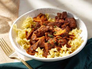 Sliced sirloin tips, halved cremini mushrooms, minced garlic, Greek yogurt, and beef broth arranged on a wooden cutting board