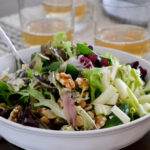 Close-up of a single serving of the October harvest salad in a white bowl, showing julienned apple ribbons, candied walnuts, and blue cheese crumbles with lemon vinaigrette drizzle