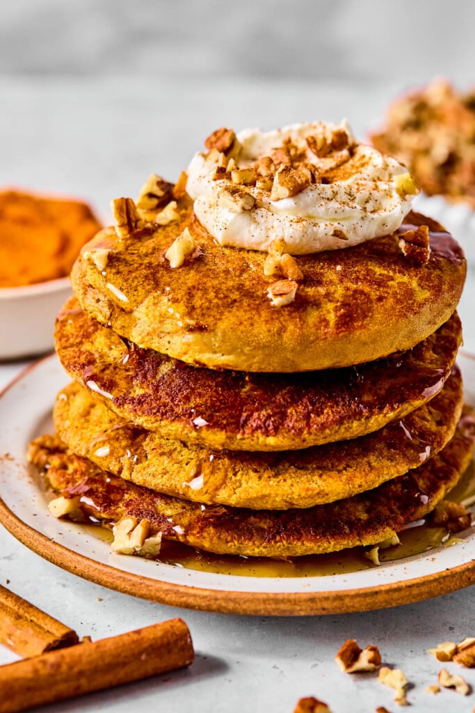 High Protein Pumpkin Pancakes — Fluffy, Nourishing & 25g+ Protein Per Stack 10 Stack of high protein pumpkin pancakes topped with Greek yogurt, a drizzle of maple syrup, and a sprinkle of pumpkin seeds on a white plate with autumn leaves in the background