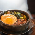 Close-up of a halved jammy egg resting on top of kimchi fried rice, the golden yolk just beginning to ooze, with visible kimchi pieces, sesame seeds, and a scattering of thinly sliced scallions