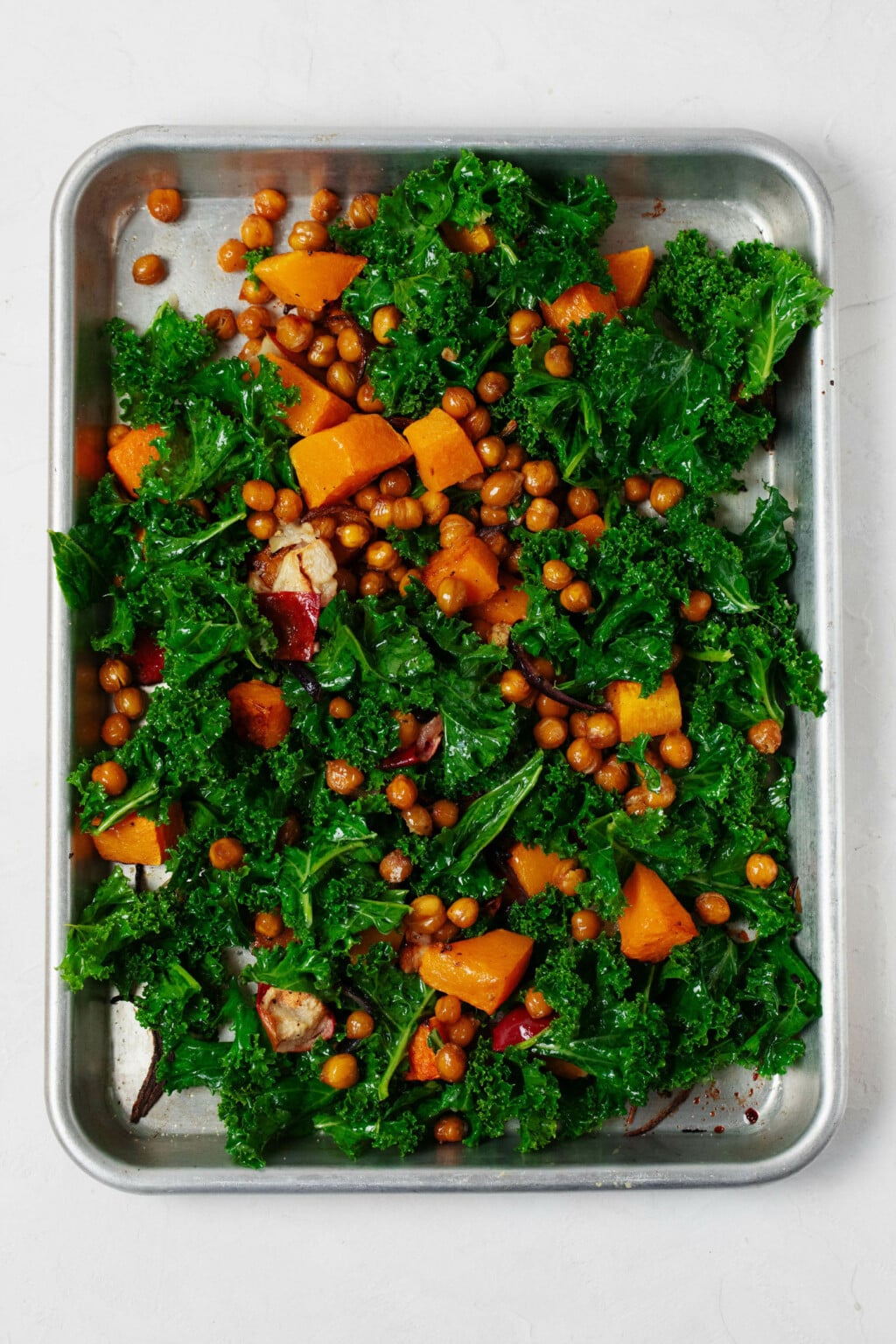 butternut squash cubes tossed in olive oil and spices on a parchment-lined sheet pan before roasting, alongside a small bowl of massaged kale and farro ingredients