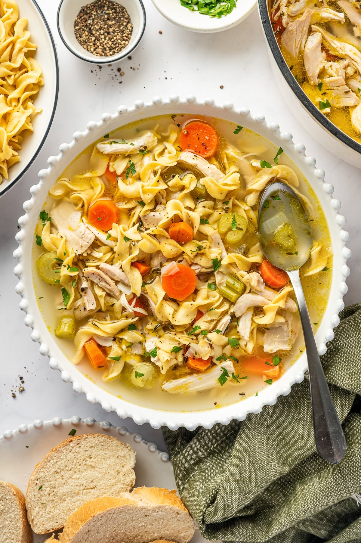 A rustic white bowl filled with lighter chicken noodle soup, loaded with whole-wheat egg noodles, tender chicken, bright carrots, and celery in a golden broth, photographed on a wooden table with fresh parsley on top