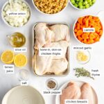 Overhead shot of lighter chicken noodle soup ingredients arranged on a clean white surface — raw chicken thighs, whole-wheat egg noodles in a bowl, halved carrots, celery stalks, garlic cloves, onion, and a carton of low-sodium chicken broth
