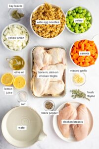 Overhead shot of lighter chicken noodle soup ingredients arranged on a clean white surface — raw chicken thighs, whole-wheat egg noodles in a bowl, halved carrots, celery stalks, garlic cloves, onion, and a carton of low-sodium chicken broth