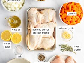 Overhead shot of lighter chicken noodle soup ingredients arranged on a clean white surface — raw chicken thighs, whole-wheat egg noodles in a bowl, halved carrots, celery stalks, garlic cloves, onion, and a carton of low-sodium chicken broth