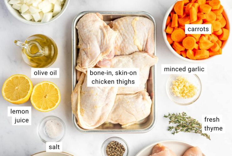 Overhead shot of lighter chicken noodle soup ingredients arranged on a clean white surface — raw chicken thighs, whole-wheat egg noodles in a bowl, halved carrots, celery stalks, garlic cloves, onion, and a carton of low-sodium chicken broth