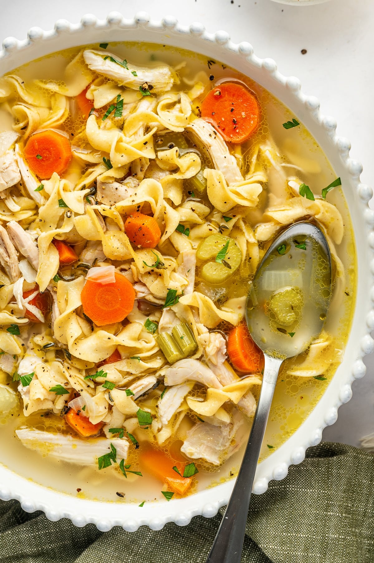Close-up of a ladle scooping lighter chicken noodle soup from a large pot, showing shredded chicken, whole-wheat noodles, and bright carrot rounds in a clear golden broth