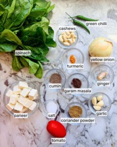 Step-by-step ingredients flat lay for Palak Paneer showing fresh spinach, paneer cubes, whole spices, ginger, garlic, and onion on a marble surface