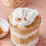 Pumpkin spice overnight oats in a glass mason jar topped with whipped cream, pumpkin seeds, and a dusting of cinnamon, photographed on a wooden board with autumn leaves in the background