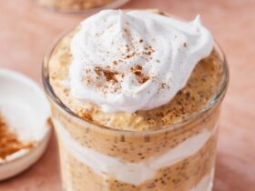 Pumpkin spice overnight oats in a glass mason jar topped with whipped cream, pumpkin seeds, and a dusting of cinnamon, photographed on a wooden board with autumn leaves in the background