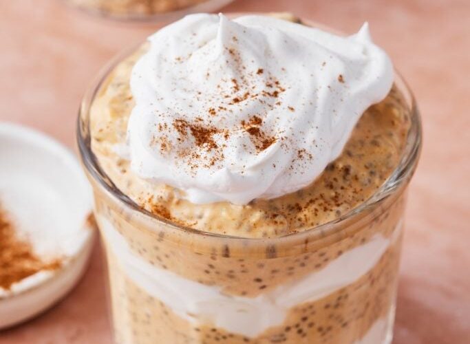 Pumpkin spice overnight oats in a glass mason jar topped with whipped cream, pumpkin seeds, and a dusting of cinnamon, photographed on a wooden board with autumn leaves in the background
