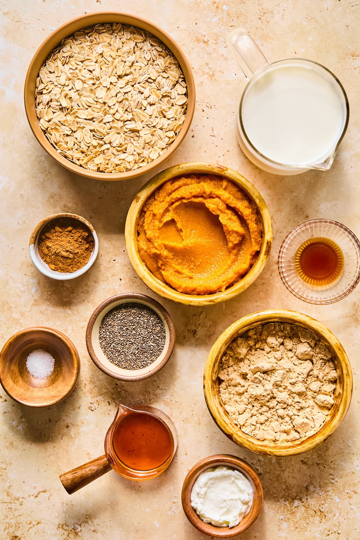 Flat lay of pumpkin overnight oats ingredients including rolled oats, Greek yogurt, canned pumpkin puree, maple syrup, vanilla protein powder, and spices arranged on a light marble surface