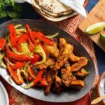 Overhead flat-lay of raw sliced chicken strips, colorful bell pepper strips, sliced onion, small bowls of fajita spices, olive oil, and fresh lime on a white marble surface