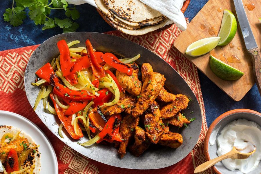 Overhead flat-lay of raw sliced chicken strips, colorful bell pepper strips, sliced onion, small bowls of fajita spices, olive oil, and fresh lime on a white marble surface