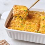 Golden tater tot casserole fresh from the oven in a white baking dish, with crispy tots on top and melted cheese bubbling at the edges, styled on a rustic wooden table