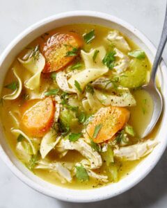 Close-up overhead shot of turkey noodle soup in a white bowl garnished with fresh parsley and cracked black pepper, with a linen napkin and crusty bread on the side
